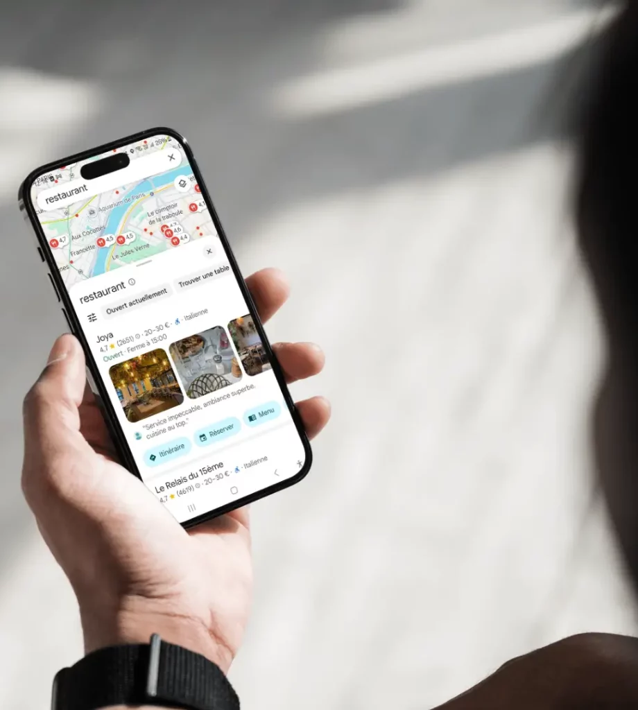 Personne tenant un smartphone affichant l’application Google Maps. L’écran montre une recherche de restaurants avec une carte en haut et une fiche de restaurant nommée « Joya » avec photos, avis et options comme « Itinéraire », « Réserver » et « Menu ».